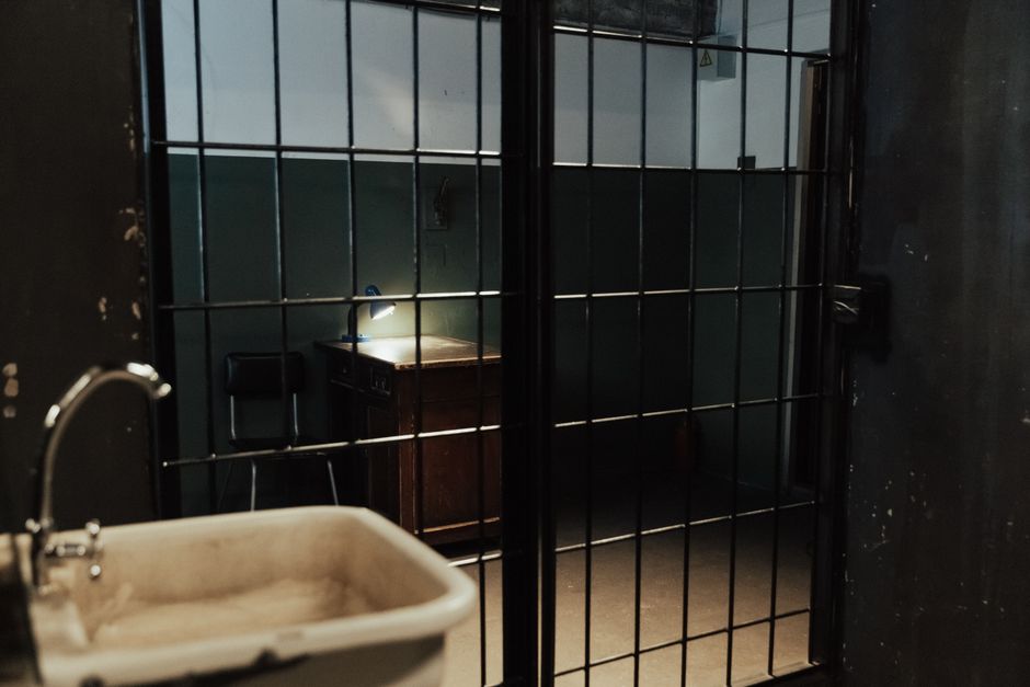 A dimly lit prison cell interior with metal bars, a sink, and a desk.
